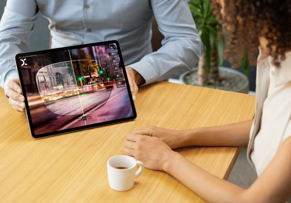 Tablet nas mãos do vendedor, virado para a cliente sentada à mesa; na tela, uma rua à noite vista pela lente dividida em "sem" e "com"